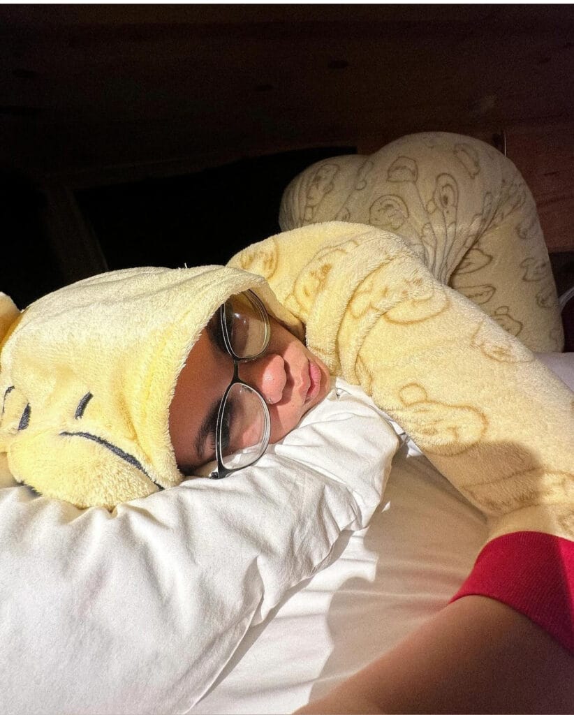 Person sleeping on a bed, wearing a yellow hooded towel with a face, and glasses resting on a white pillowglove scene under dim lighting