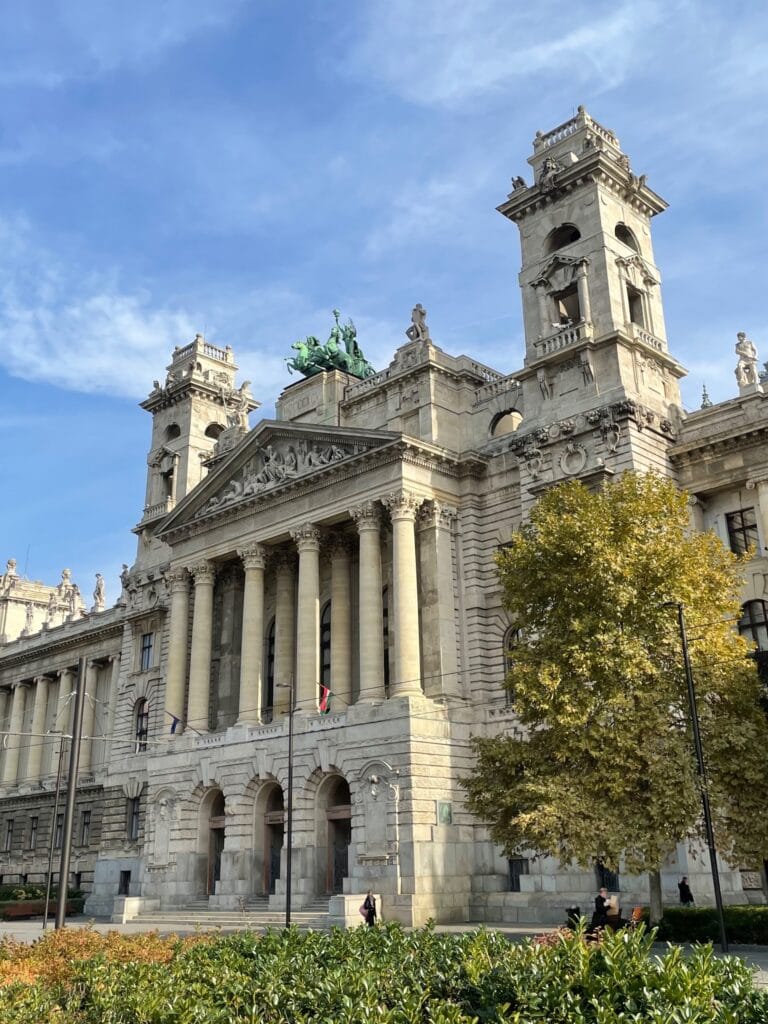 Grand stone historic building with tall towers, a colonnaded façade, and statues along the roofline against a blue sky.