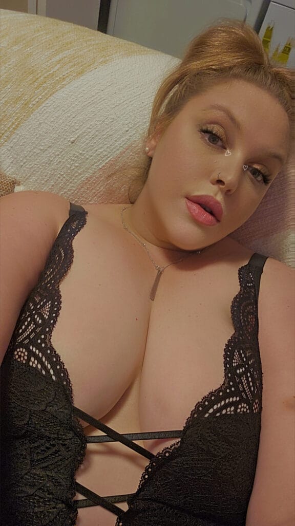 Woman in black lace lingerie lying on a beige sofa, looking at the camera with makeup and jewelry visible.