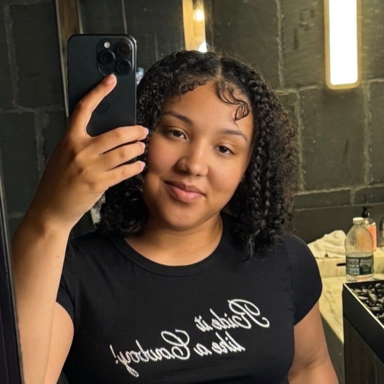 Young woman takes a mirror selfie with a smartphone, wearing a black T-shirt in a backstage/dressing-room setting with warm lighting.