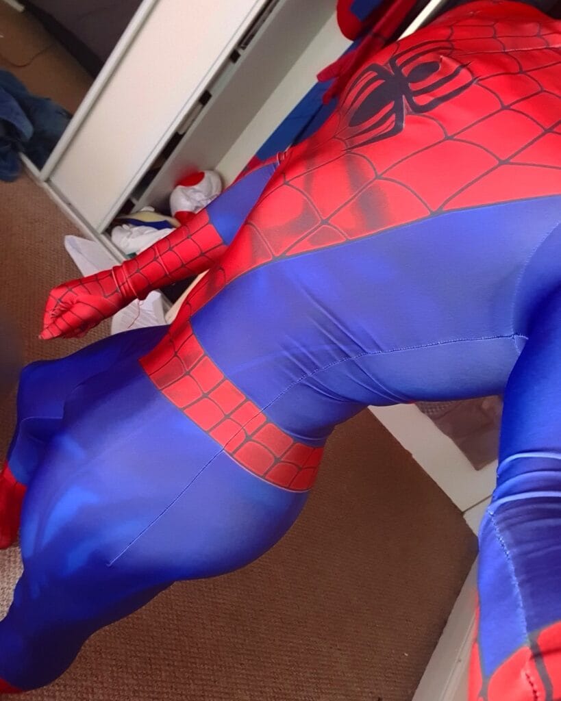 Child wearing a Spider-Man blue and red costume sitting on a bed with a cluttered room in the background.