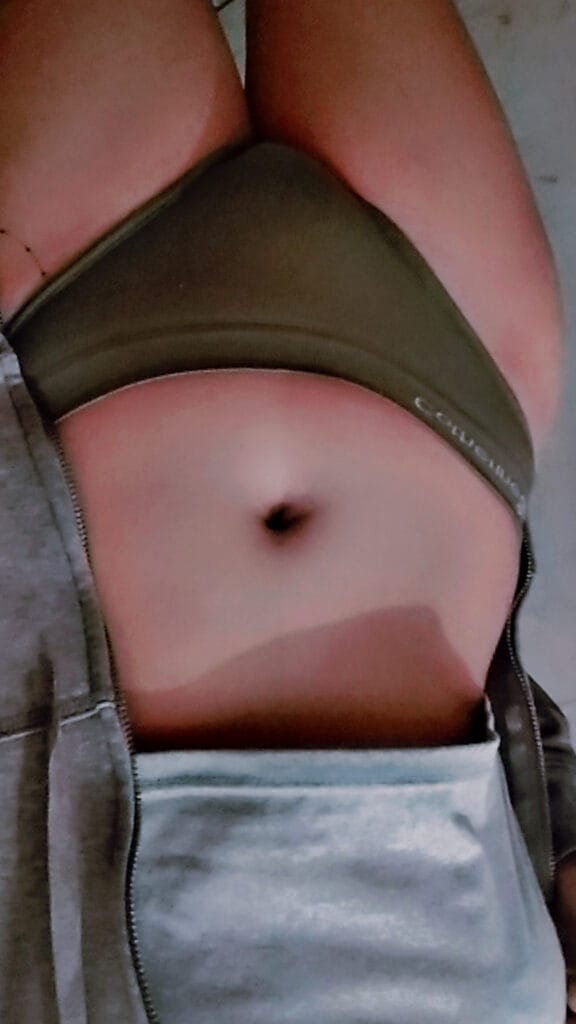 Close-up of a person wearing a green sports bra and gray zip-up hoodie, showing the midsection and belly button; the face is not visible.