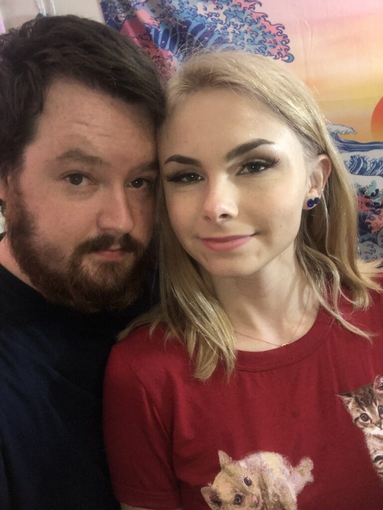 A man with dark hair and beard and a smiling blonde woman in a red shirt posing for a selfie, with a colorful wave mural in the background.