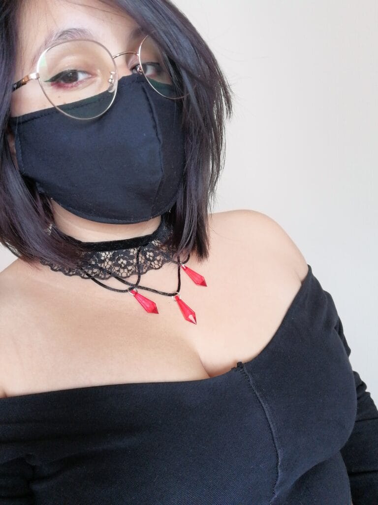 Person wearing a black face mask, round glasses, and a black off-shoulder top with lace choker and red triangular earrings.
