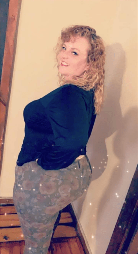 Smiling woman with curly blonde hair standing sideways against a beige wall, wearing a black top and camo-patterned pants, with sparkles in the foreground.