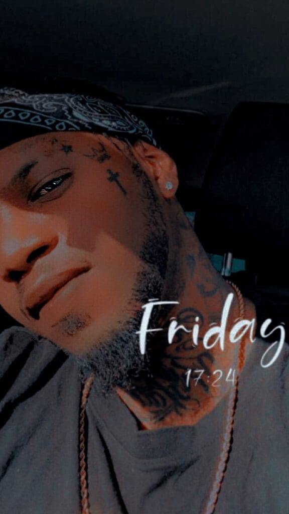 Close-up portrait of a man wearing a blue bandana, visible facial tattoos, an earring and a necklace, inside a car with the word 'Friday' overlaid on the right.
