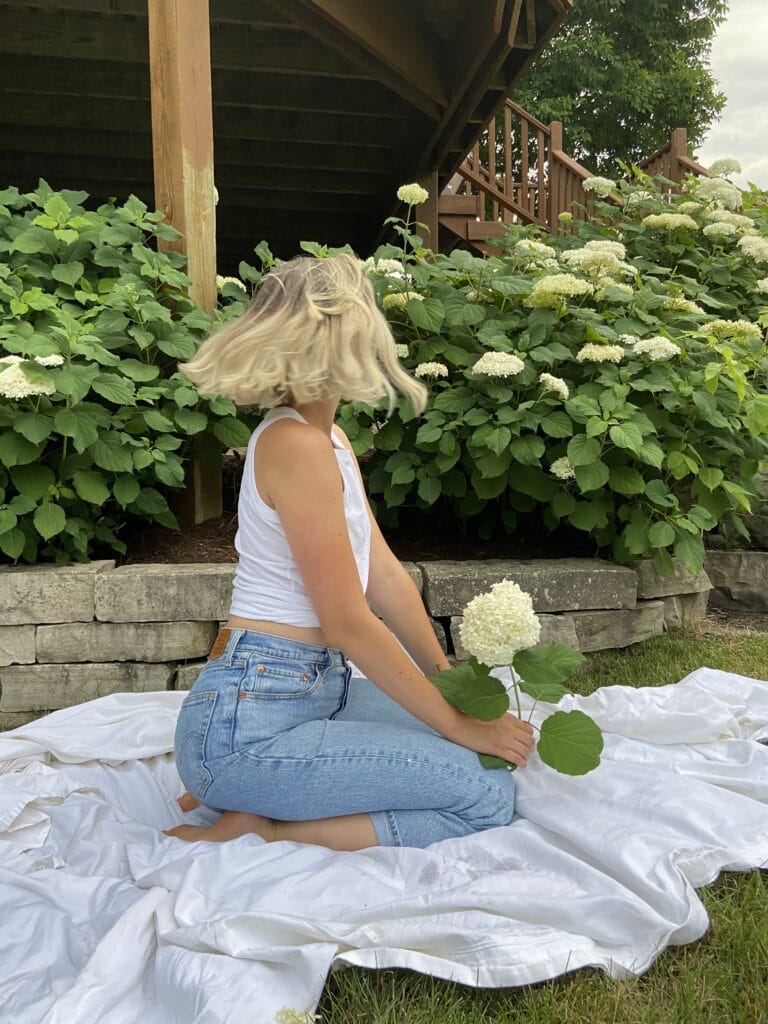 Woman kneeling on a white blanket beside a hydrangea bush with large white blossoms under a wooden deck structure.