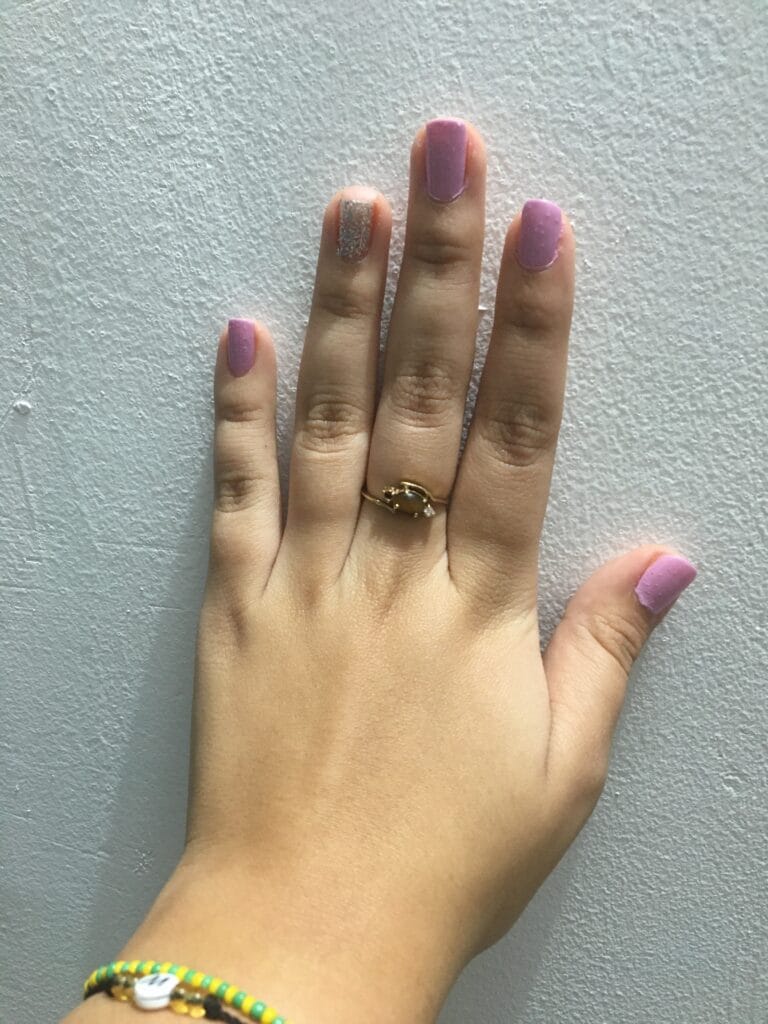 Hand showing lavender-pink nails with one glitter accent nail, wearing a gold ring and a colorful bracelet against a textured gray wall.
