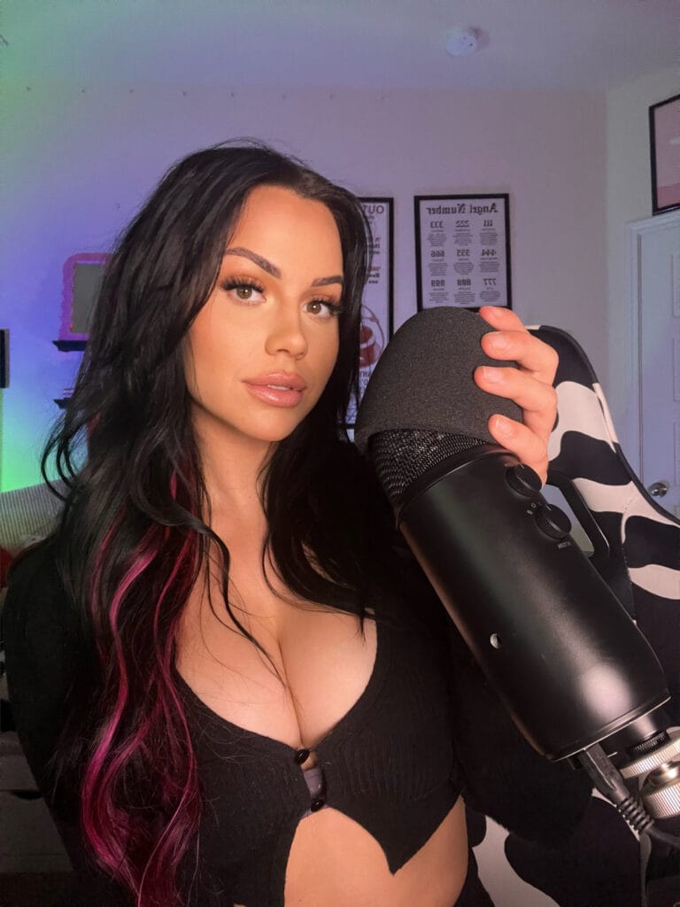 Close-up portrait of a woman with dark hair and pink streaks holding a microphone, bathed in colorful room lighting.