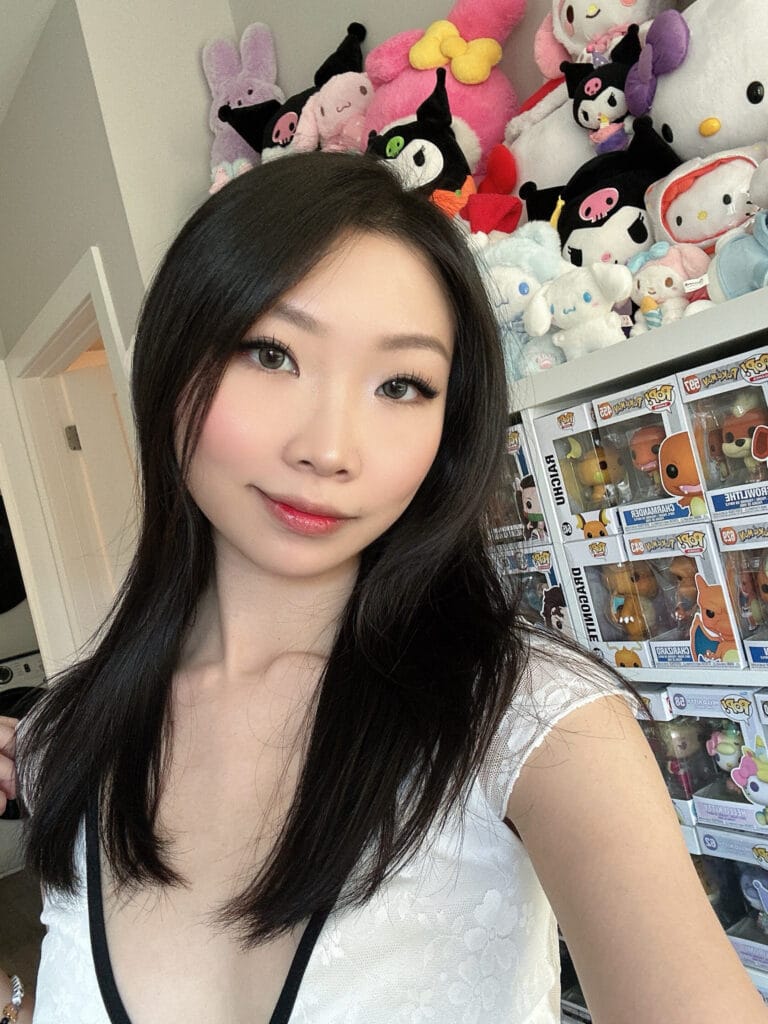 Portrait of a smiling woman with long dark hair taking a selfie, plush toys on a shelf behind her.
