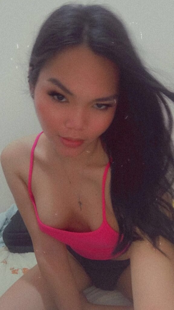 Close-up selfie of a woman with long dark hair wearing a bright pink bikini top, looking at the camera with a neutral expression in a bedroom setting.