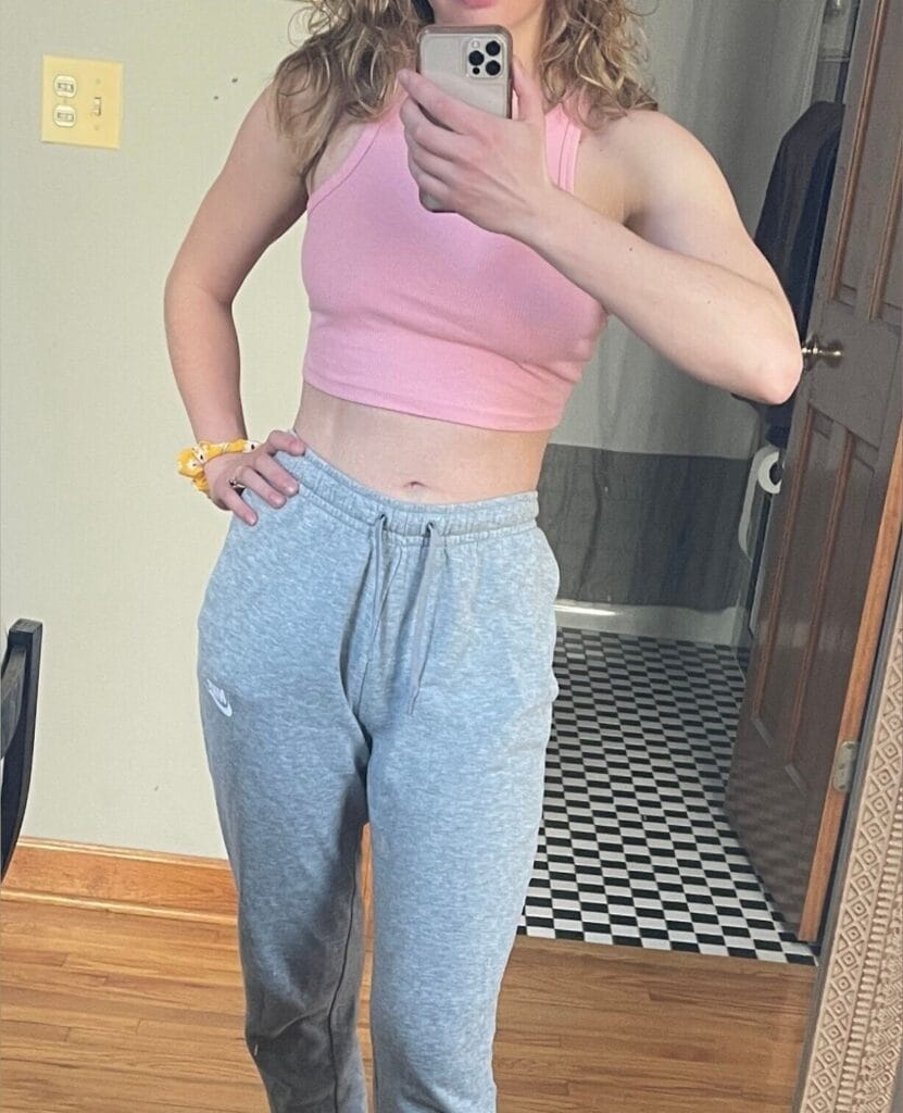 Person taking a mirror selfie in a bathroom, wearing a pink cropped tank top and gray sweatpants, holding a phone in front of their face.