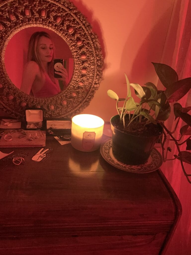 A woman taking a mirror selfie in a round woven-frame mirror with a candle, plant, and jewelry on a wooden dresser bathed in red light