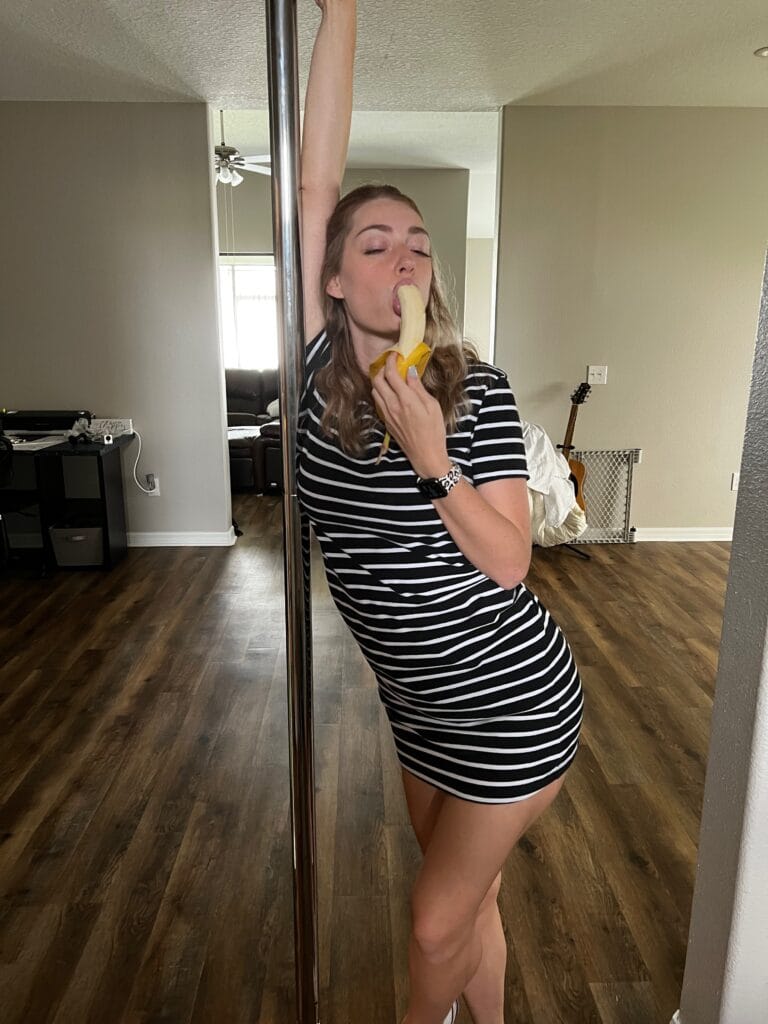 Woman in a black-and-white striped dress posing with a vertical pole, one arm raised, indoors.