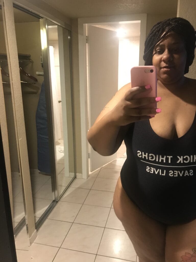 Woman taking a mirror selfie in a bathroom, holding a pink phone and showing pink nails.